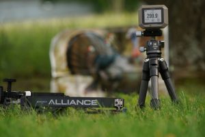 Speed - North American Bow Hunter Tripod-mounted device with a digital readout in a grassy outdoor setting. The screen shows numbers and is in focus, with blurred background equipment.