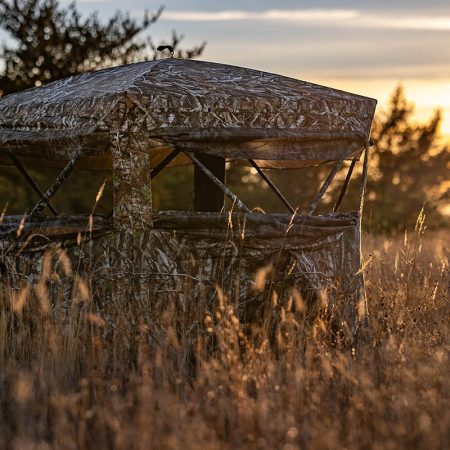 optivue-3600-ultra-hd-see-through-hunting-blind-with-900-full-access-door-kol-exclusive-169005_d281e410-a430-4405-8fdd-f2afb3667661_1200x - North American Bow Hunter Camouflage hunting blind in a field of tall grasses at sunset, with a wooden frame and mesh walls.