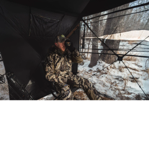 blind22 - North American Bow Hunter Hunter in camouflage seated inside a ground blind, looking through binoculars at a snowy forest outside.