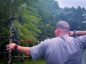 feature ryan - North American Bow Hunter Man wearing a backward cap and gray shirt draws a compound bow outdoors among green trees, aiming with a sighted bow.