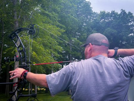 feature ryan - North American Bow Hunter Man wearing a backward cap and gray shirt draws a compound bow outdoors among green trees, aiming with a sighted bow.