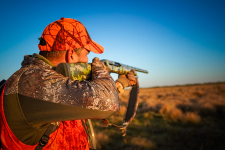ammo feat - North American Bow Hunter Hunting scene: hunter in camo jacket and orange cap aiming a scoped rifle across a dry field.